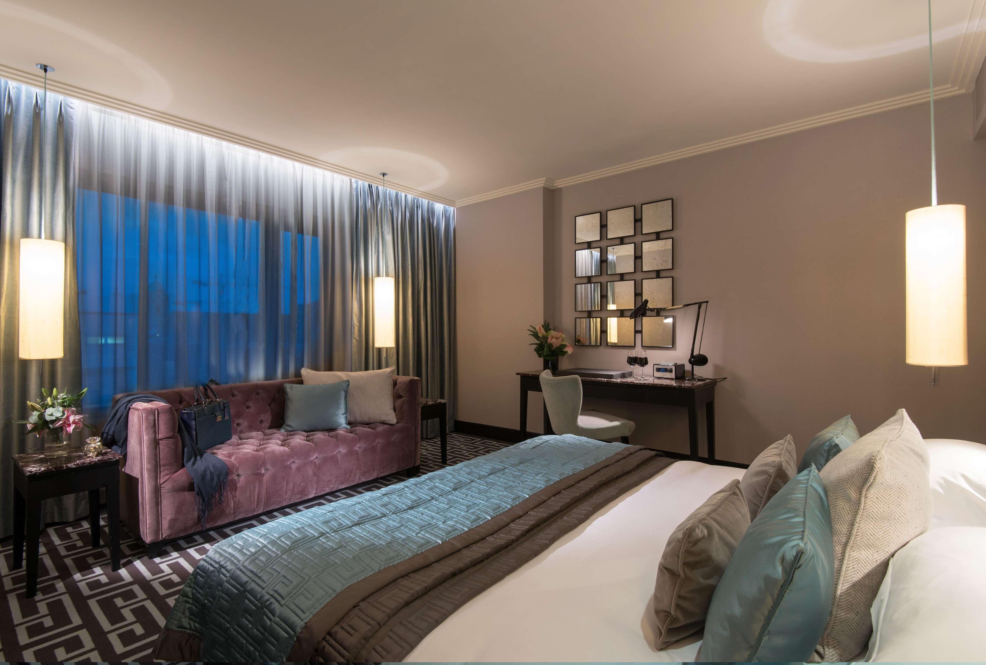 The Fitzwilliam Hotel Belfast Exterior foto The photo depicts a modern hotel room featuring a spacious layout. There is a large bed with decorative pillows in the foreground, covered with a stylish bedspread. To one side, a plush purple sofa adds a pop of color and comfort. A small desk and ch